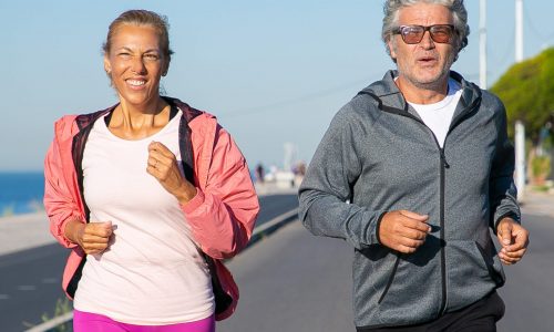 Cheerful mature couple running along river bank. Grey haired man and woman wearing sports clothes, jogging outside. Active lifestyle and age concept