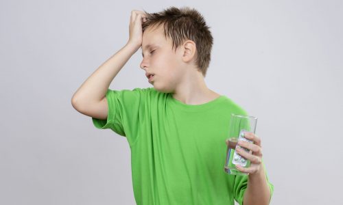 ill little boy in green t-shirt feeling unwell holding glass of water and pills touching his head suffering from cold standing over white background