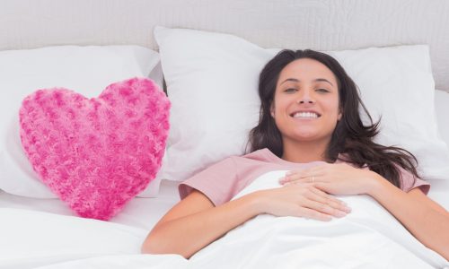 Woman lying in her bed next to a pink heart pillow and smiling at camera